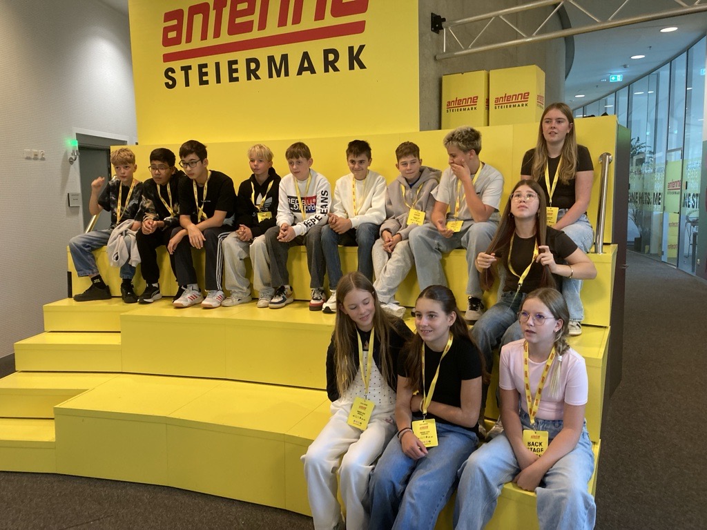 Kinder und Jugendliche sitzen auf gelber Bühne vor Antenne Steiermark Logo